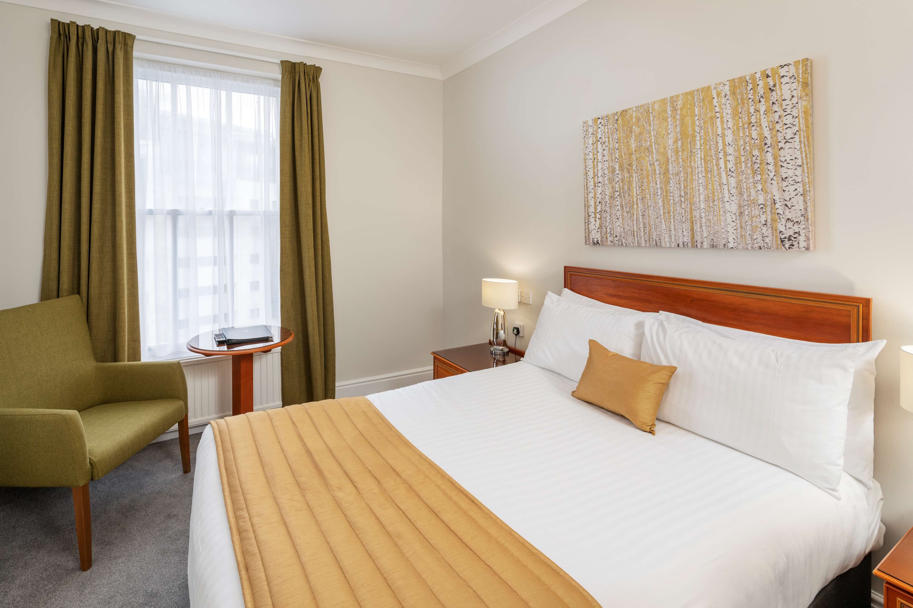 A hotel room with a bed, a green armchair, a small table with a lamp, and a painting on the wall.