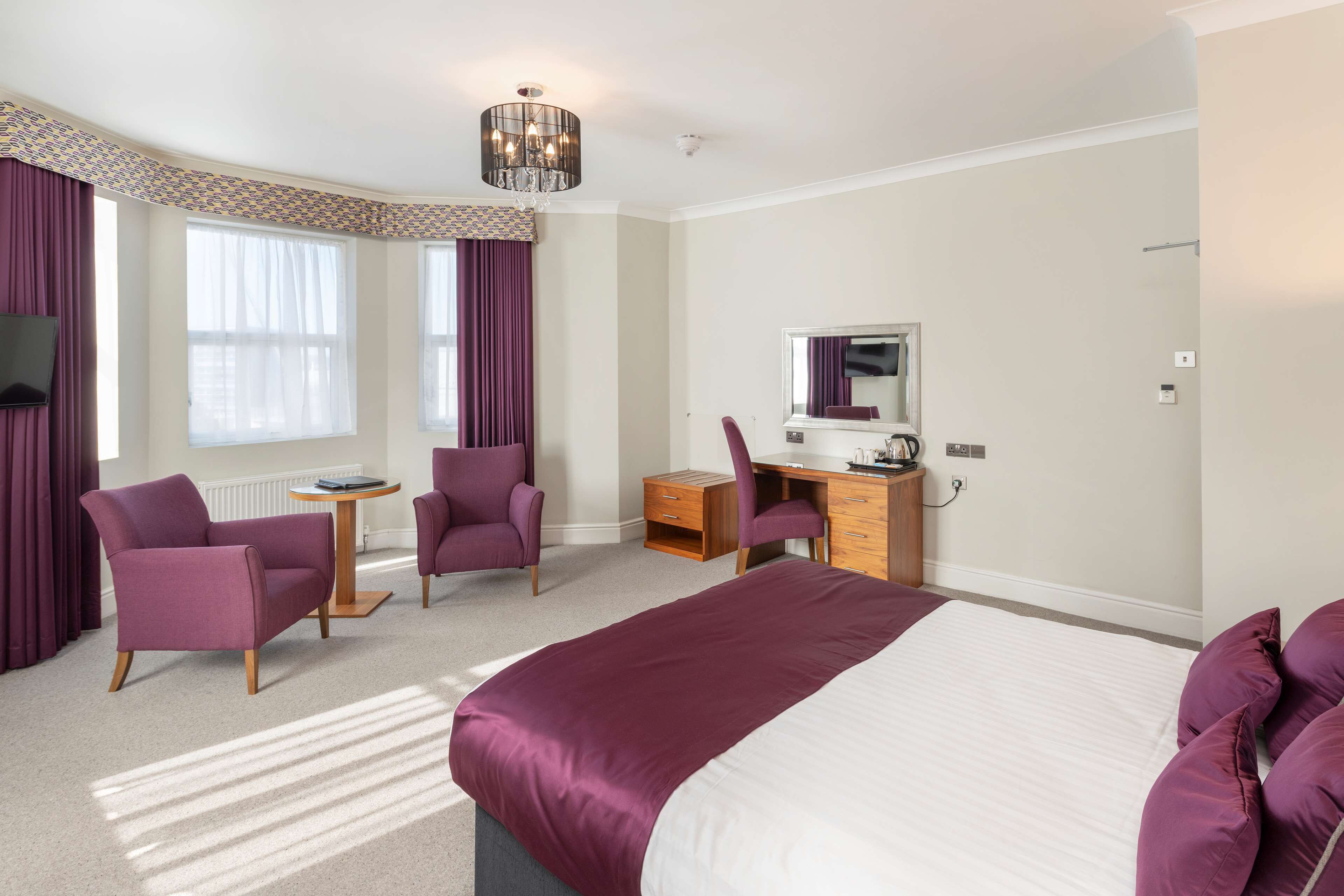 Une chambre d’hôtel avec un grand lit, deux fauteuils violets, un bureau en bois, une petite table ronde et un grand miroir.