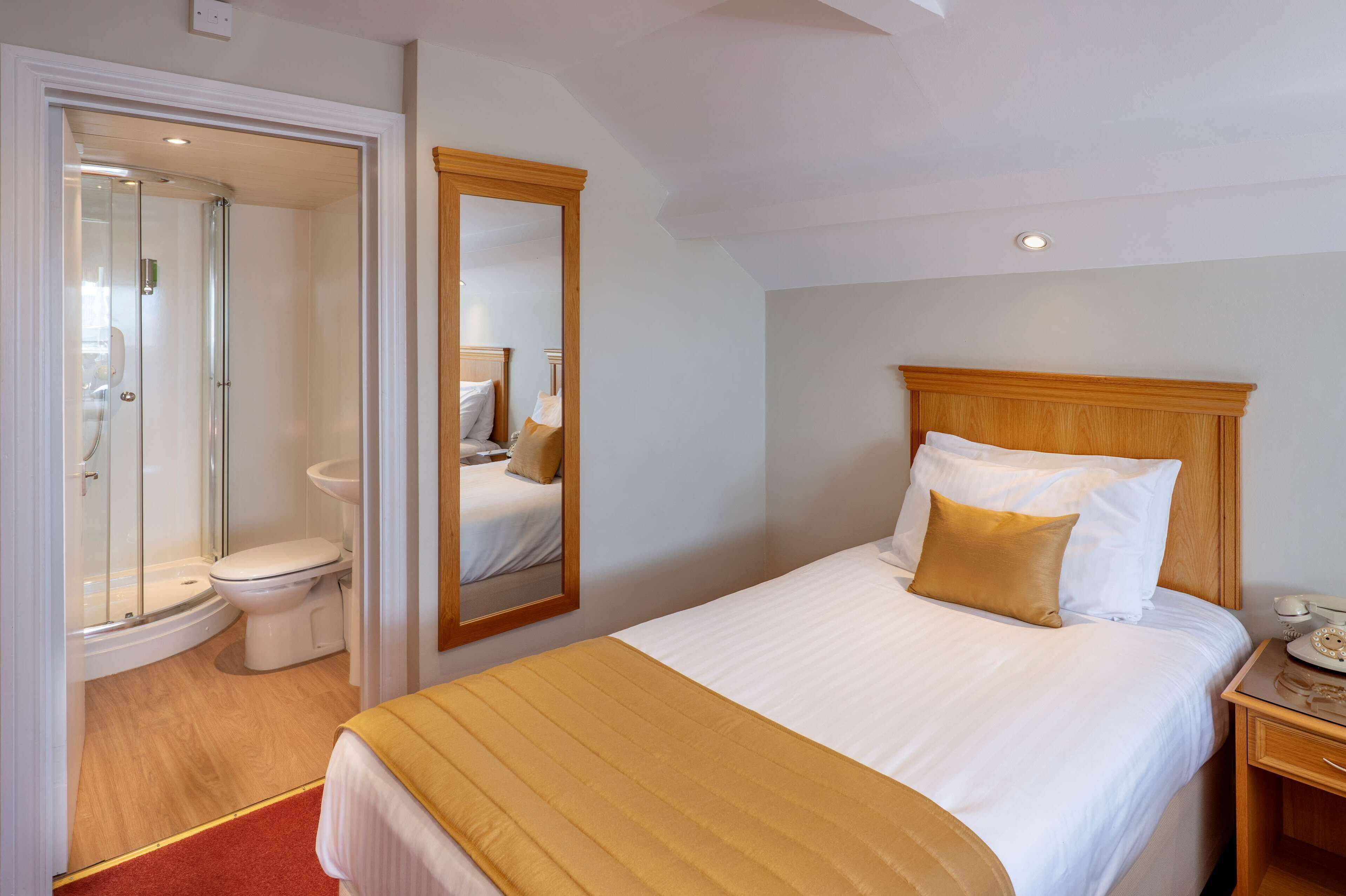 Una habitación de hotel con una cama, una mesita de noche, un espejo, un cabecero de madera, ducha y un inodoro.