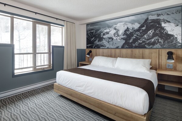 A hotel room with a large bed, a window with curtains, a wall-mounted lamp, and a mountain landscape painting.