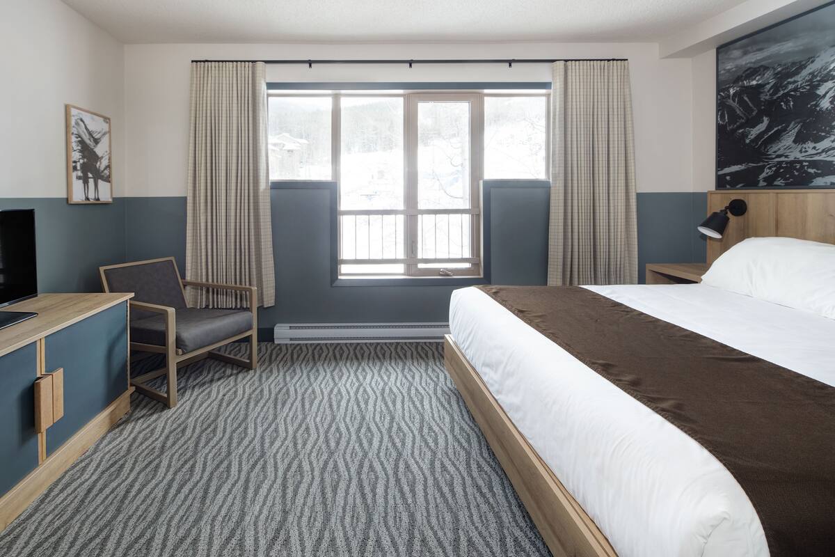 A hotel room with a bed, a desk, a chair, a window with curtains, and a painting on the wall.