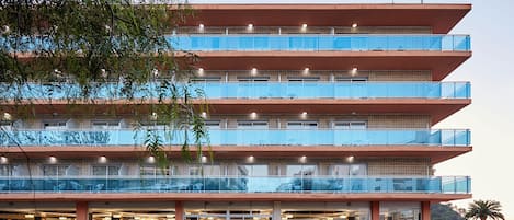 Ein mehrstöckiges Hotel mit einem Glasbalkon in den oberen Etagen, einem Restaurant im Erdgeschoss mit Außenbereich und einem Pool mit einem markanten Design.