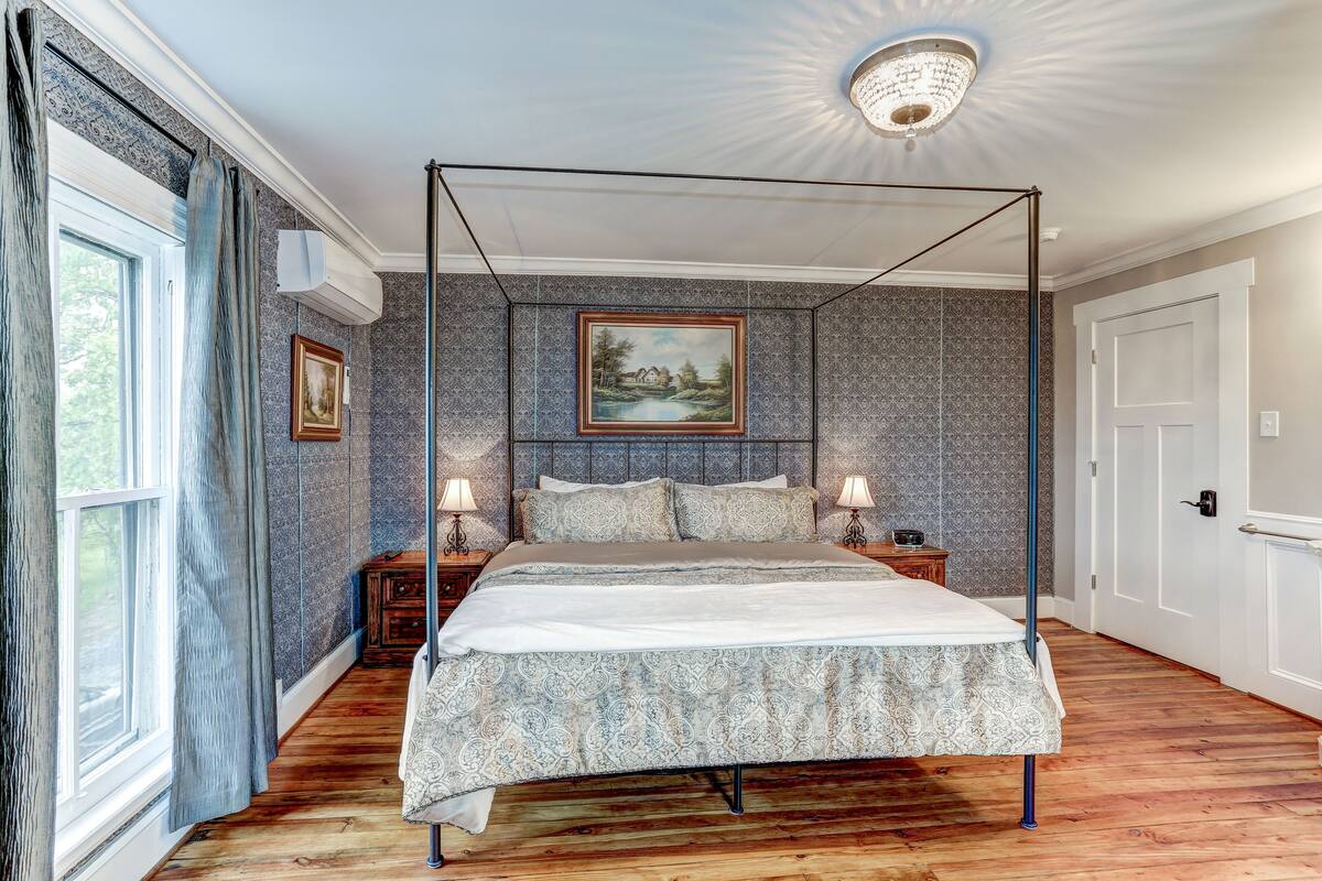 A four-poster bed with a canopy, a painting above it, and a view of the outdoors through a window.