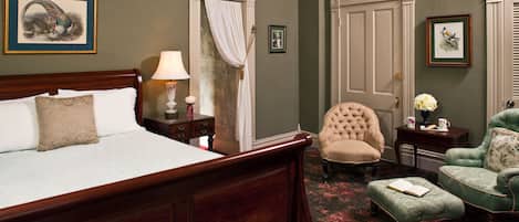 A bedroom with a large wooden bed, a green armchair, a matching ottoman, a nightstand with a lamp, and a framed picture on the wall.