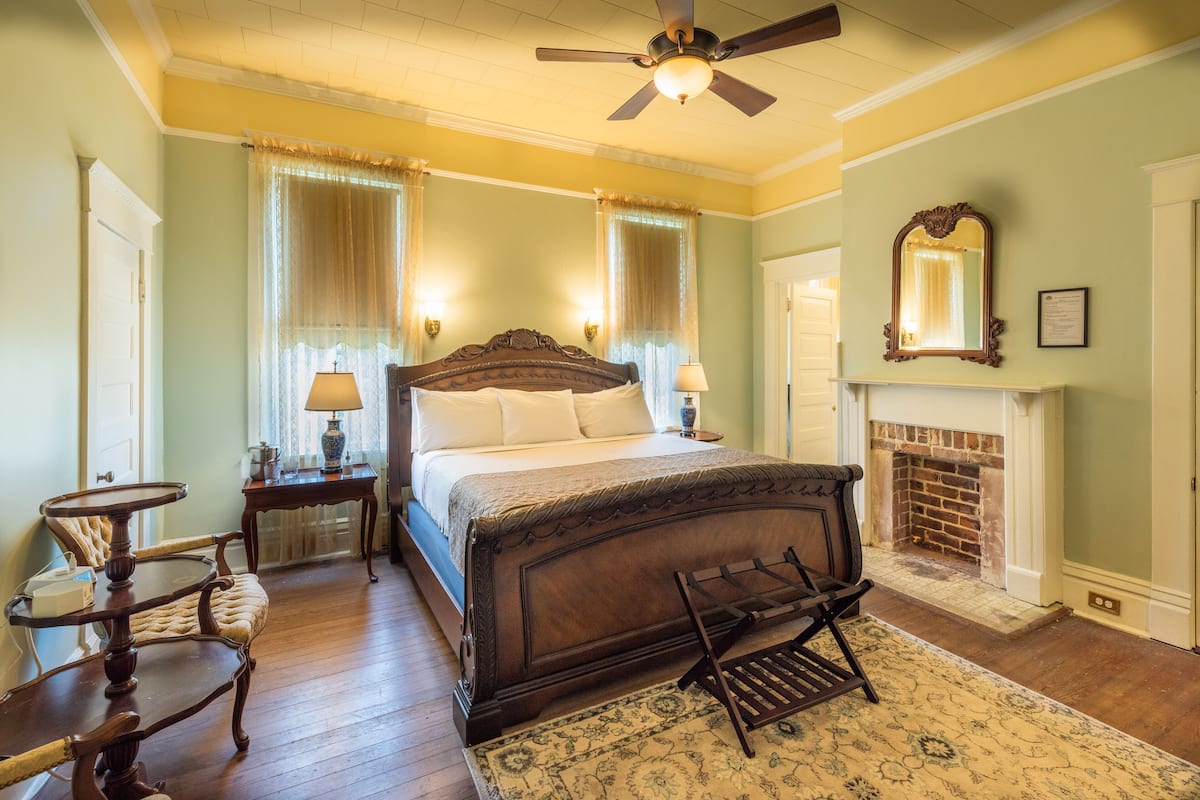 A bedroom with a large bed, a wooden chair, a small table, a fireplace, and a ceiling fan.