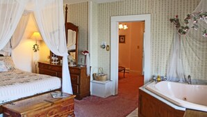 Un dormitorio con una cama con dosel, un aparador de madera, una tocador con espejo, una bañera grande y una puerta que da a otra habitación.