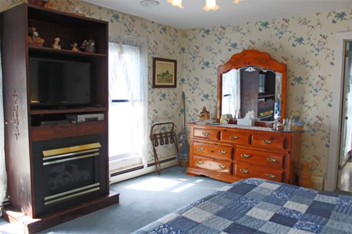Un dormitorio con un aparador de madera, televisor, colcha acolchada, papel tapiz floral y un cuadro en la pared.