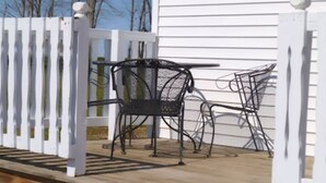 Una terraza de madera con sillas de metal y una mesa, delimitada por una valla de piquetes blancos.