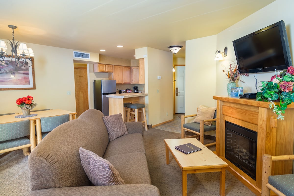 A cozy living room with a sofa, a coffee table, and a fireplace. There is a kitchen area with cabinets and a refrigerator. A television is mounted on the wall.