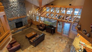Amplia sala de estar con chimenea de piedra, muebles de madera y ventanas grandes que ofrecen vistas del interior.