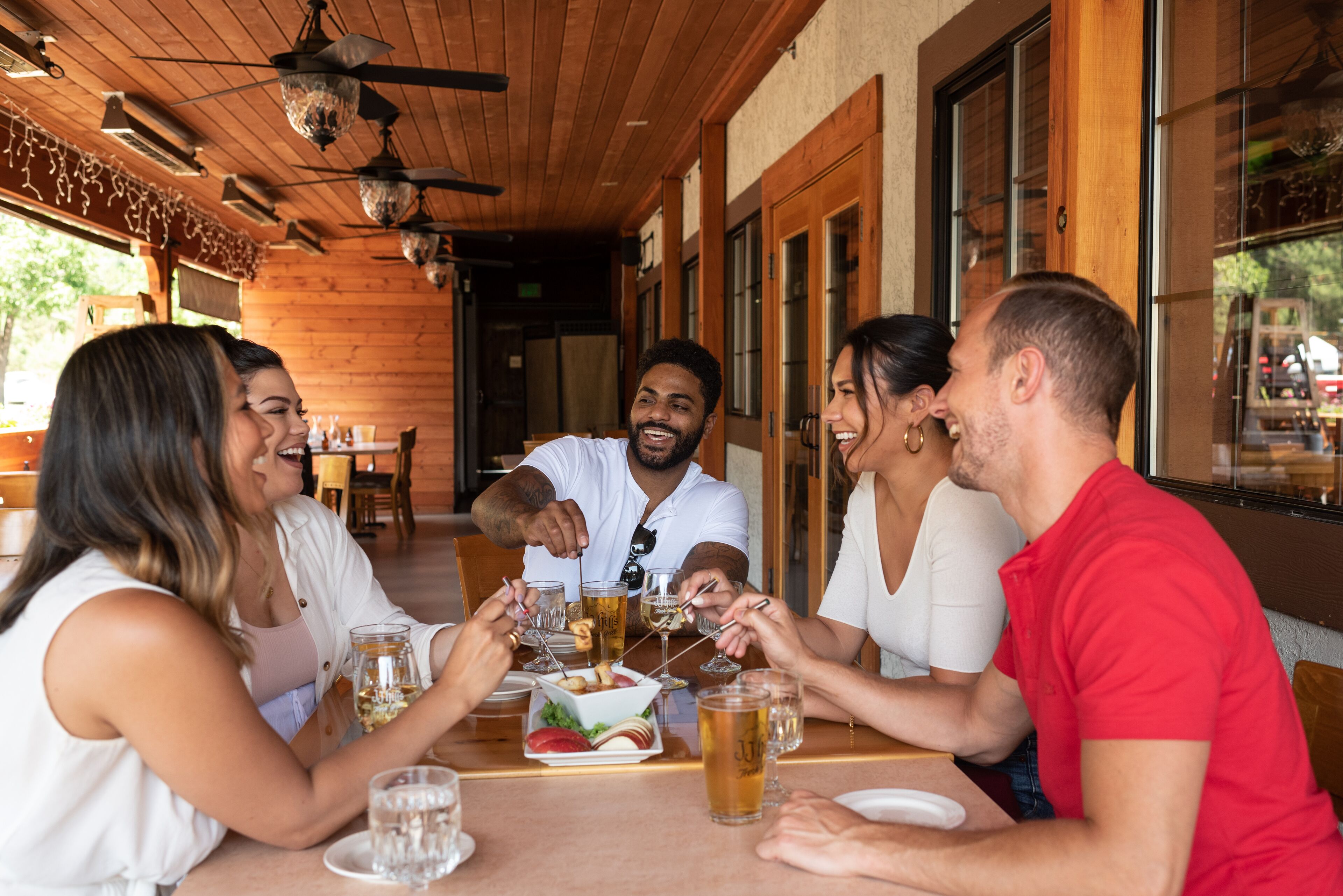 A group of people toasting at a wooden table with drinks and snacks in a restaurant with a wooden interior and large windows.