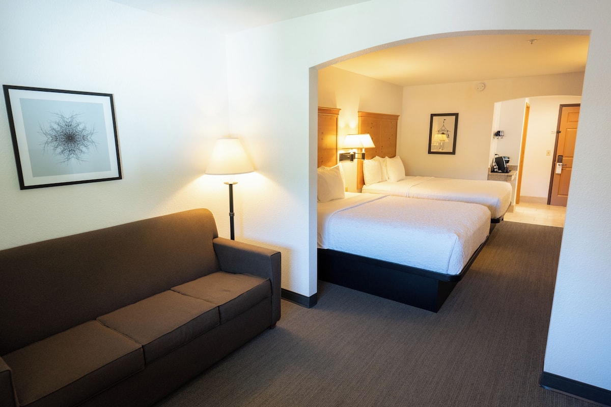 A hotel room with a sofa, two beds, a lamp, and a framed picture on the wall.