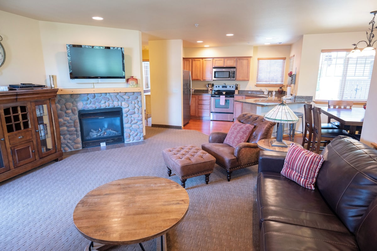 A living room with a fireplace, a television mounted above it, a sofa, a coffee table, and a kitchen area in the background.