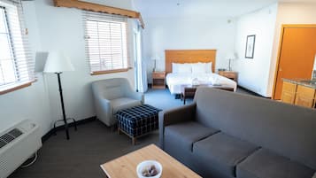 Une chambre d’hôtel comprenant un lit, deux canapés, une table avec un saladier, une chaise, un bureau, une lampe, un radiateur et une fenêtre avec des stores.