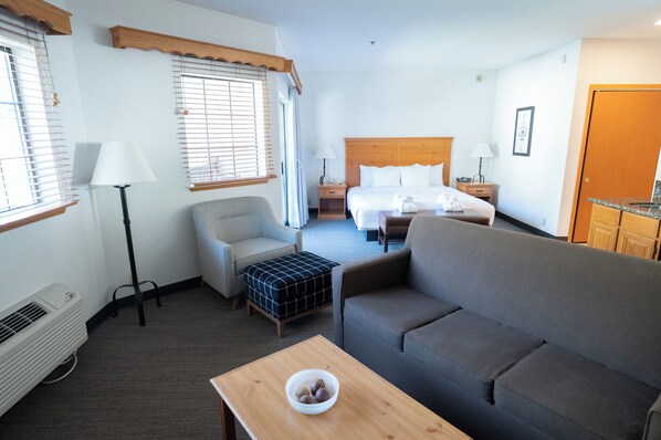 Habitación de hotel con cama, dos sofás, una mesa con un tazón, una silla, un escritorio, una lámpara, un calentador y una ventana con persianas.