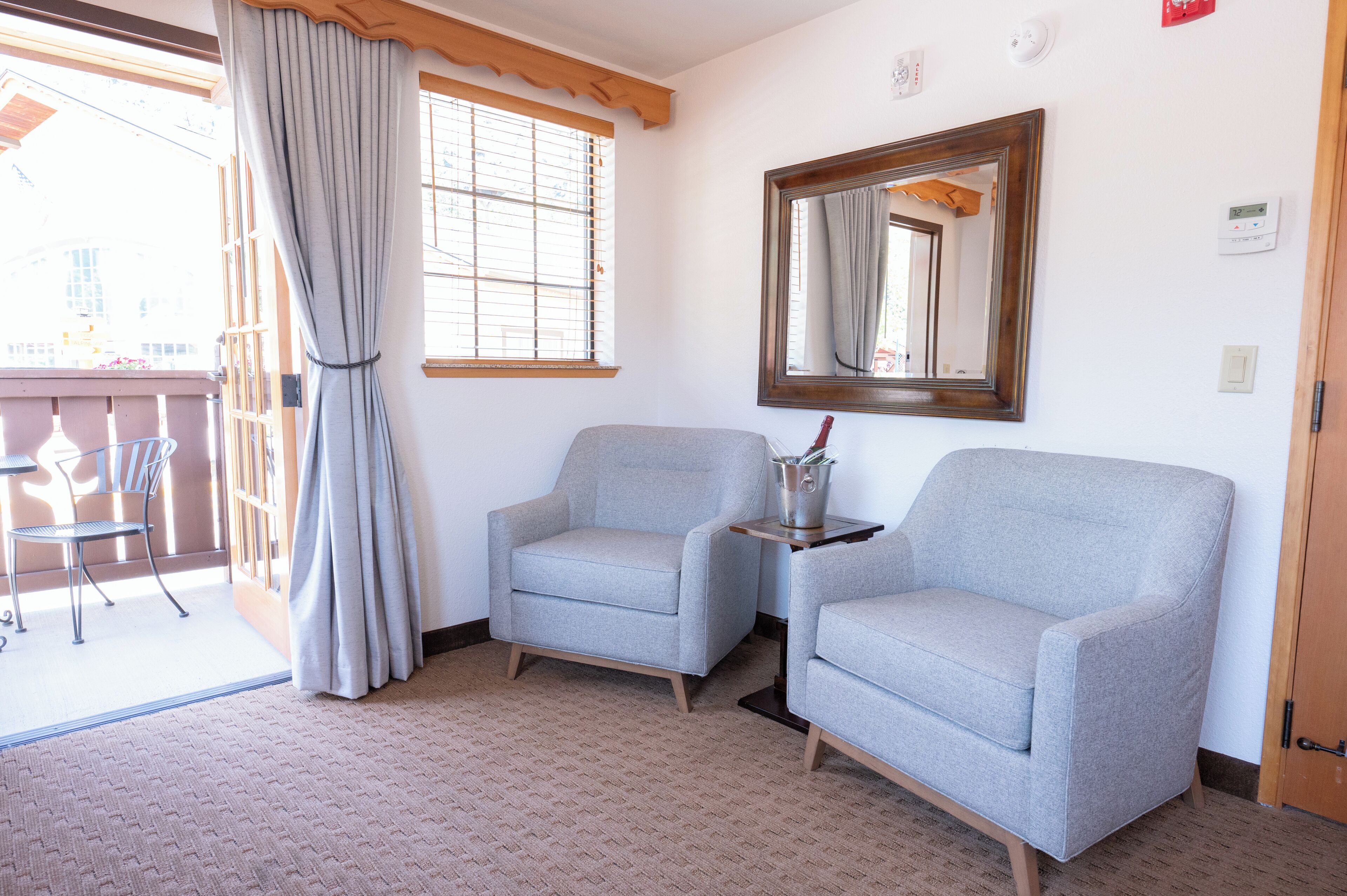A room with two grey armchairs, a small table with a metal bucket, a window with curtains, and a wooden door.
