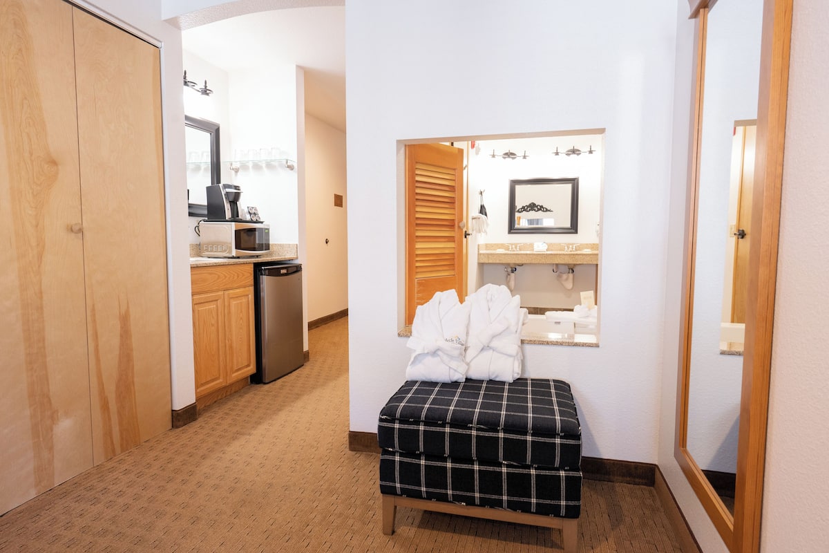A hotel room with a kitchenette, a bathroom with a mirror and towel rack, and a bench with towels.