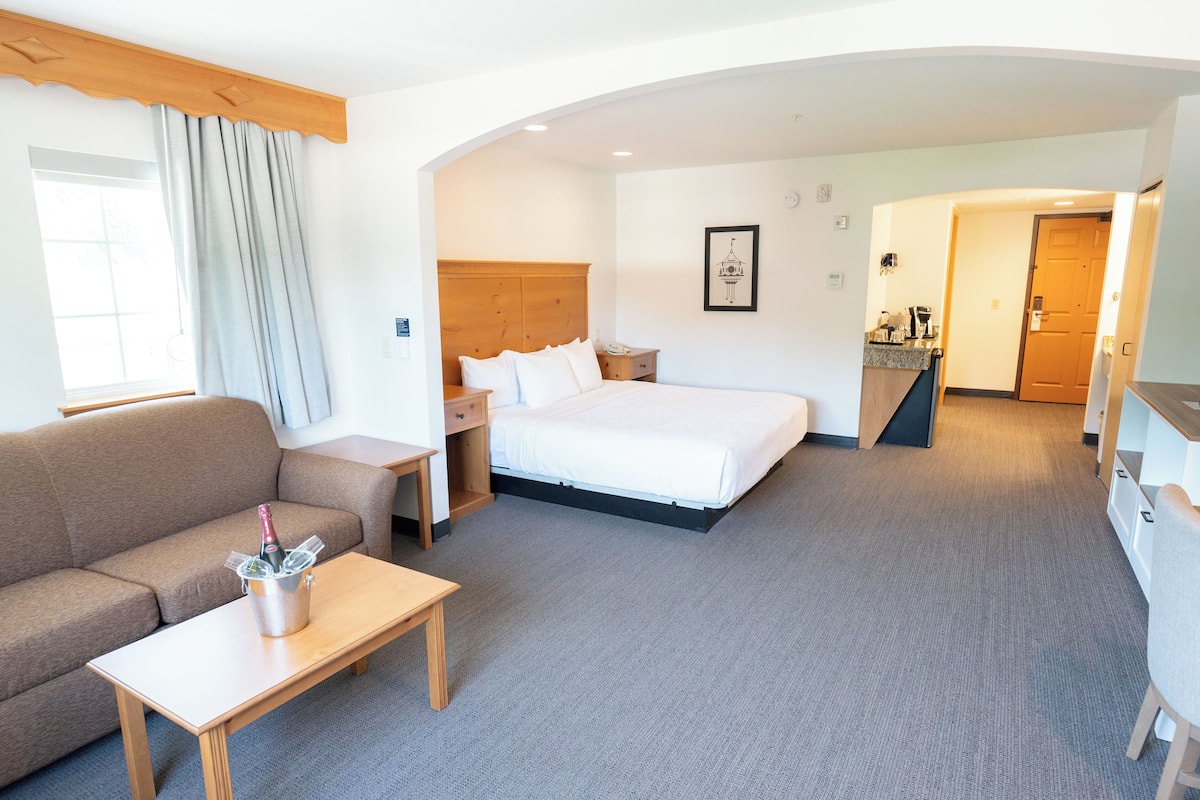 A hotel room with a bed, a sofa, a small table with a bottle, a wooden headboard, and a framed picture on the wall.