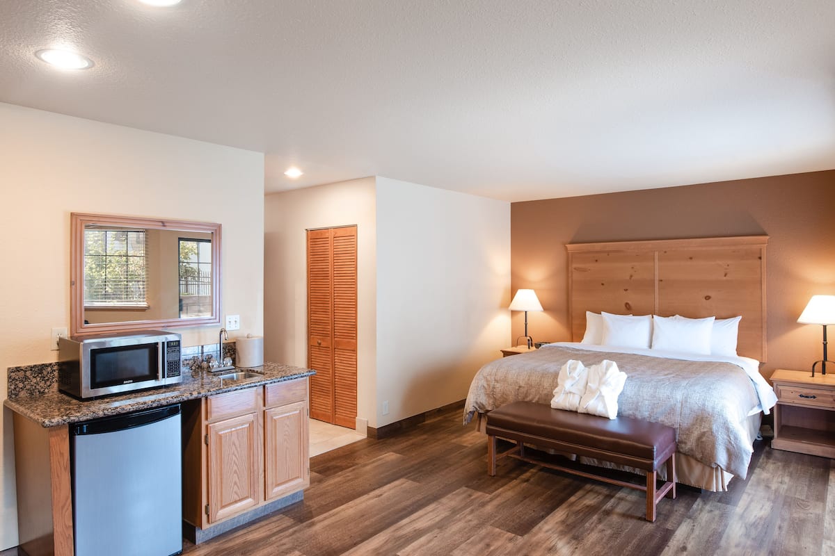 A hotel room with a kitchenette, a bed with a wooden headboard, a bench, and a nightstand with a lamp.