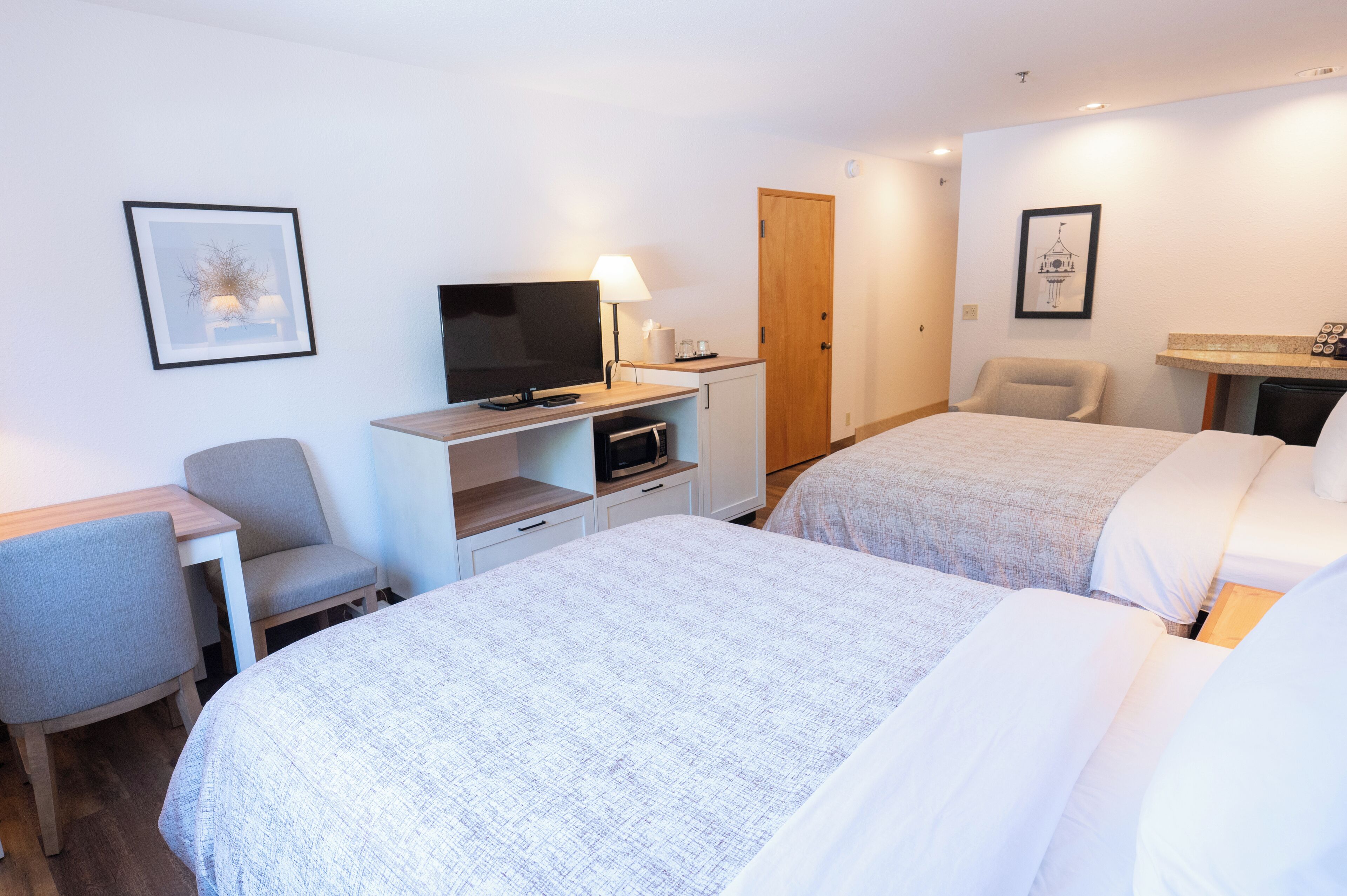 A hotel room with two beds, a desk with a TV, a chair, and a small table with a lamp.