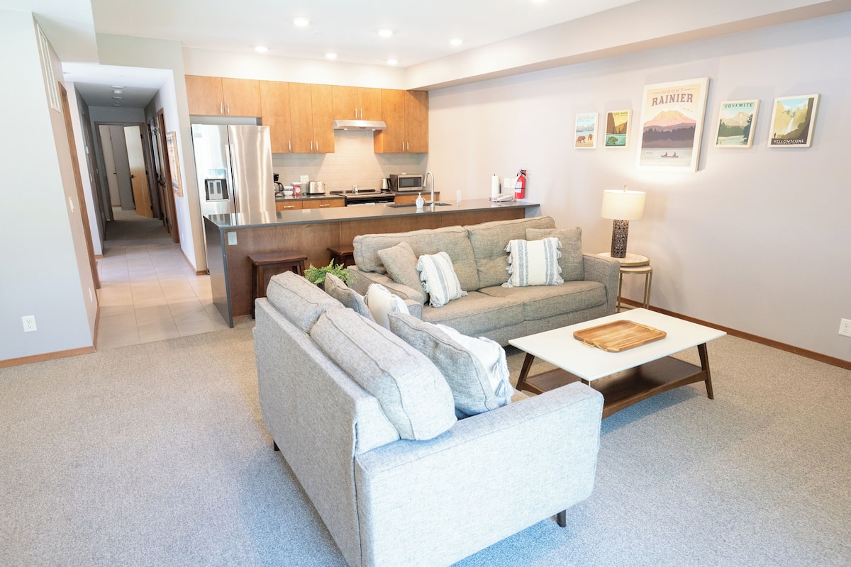 A modern living room with a grey sectional sofa, a white coffee table, and a kitchen area with wooden cabinets and stainless steel appliances.