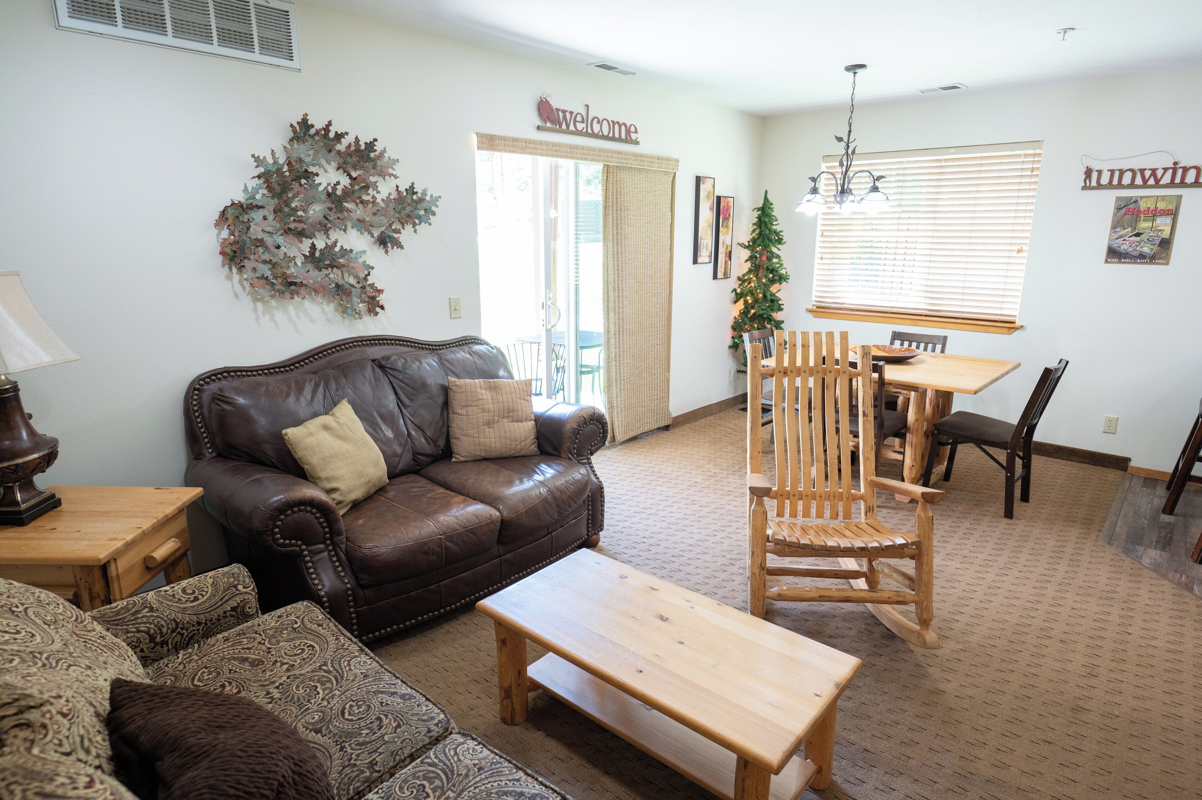 A living room with a brown leather sofa, a wooden coffee table, a patterned armchair, and a wooden rocking chair. There is a dining table with chairs, a Christmas tree, and a 'Welcome' sign above the door.