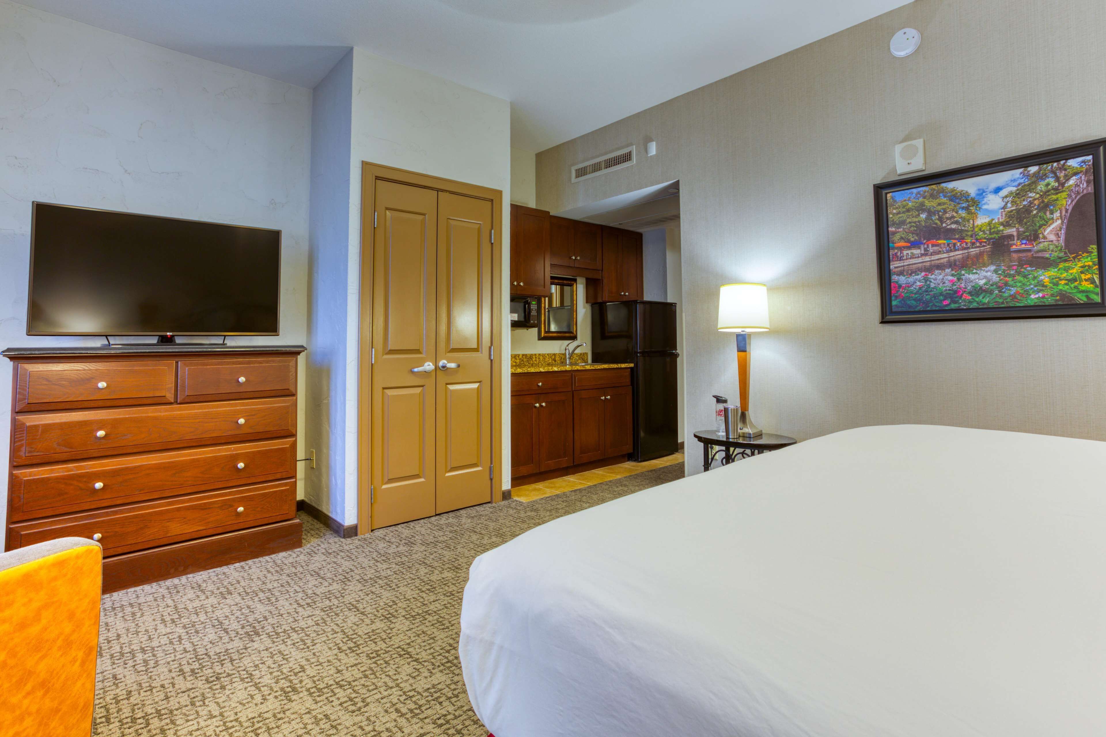 A hotel room with a bed, a TV, a dresser, a kitchen area, and a painting on the wall.