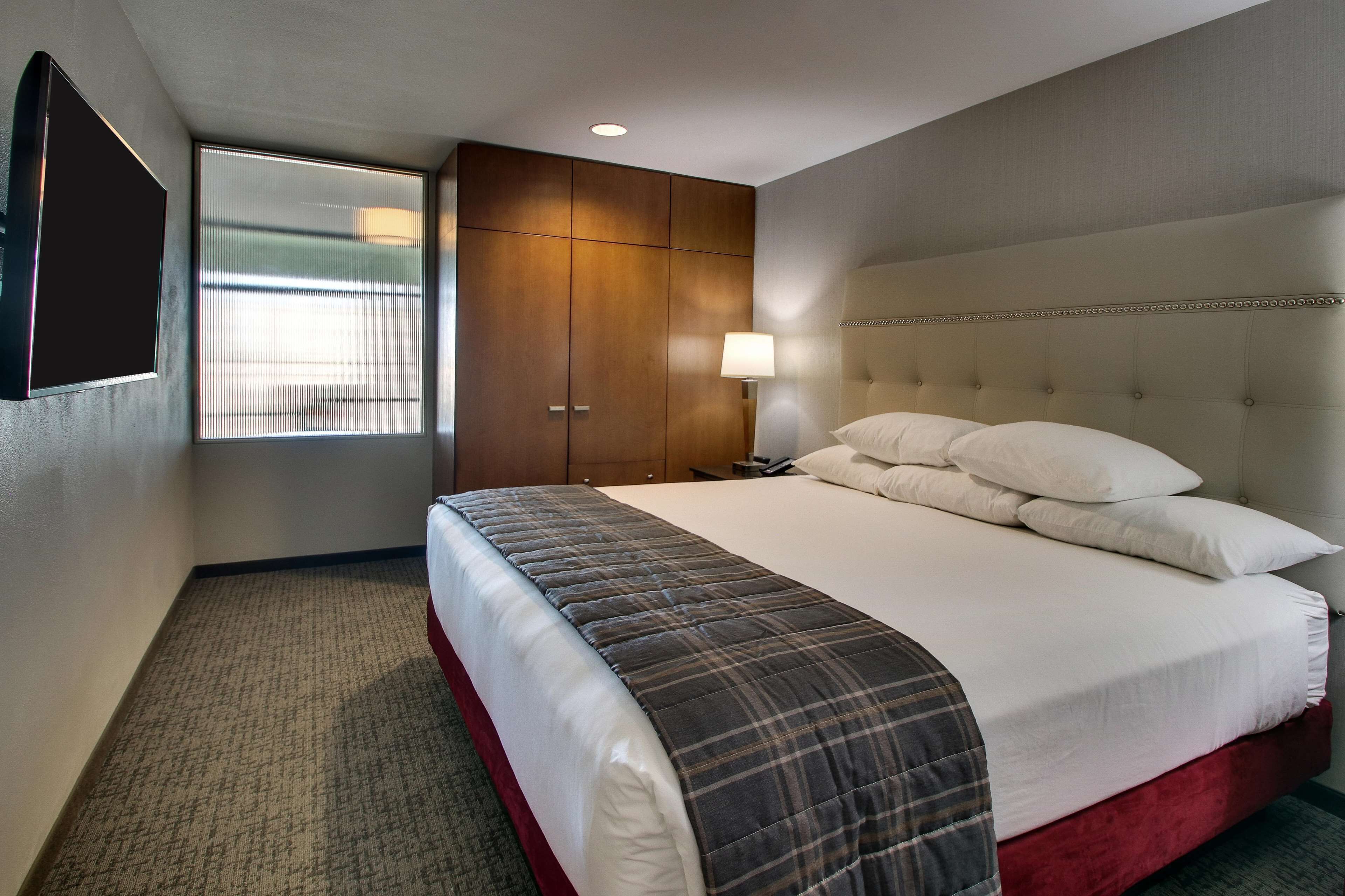 A hotel room with a large bed, a wardrobe, a bedside table with a lamp, and a window with blinds.
