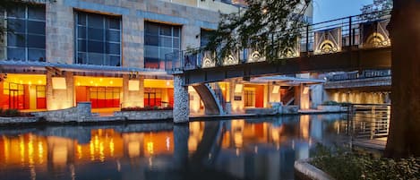 A modern hotel with a stone facade, a reflective water feature, and a bridge connecting different sections.