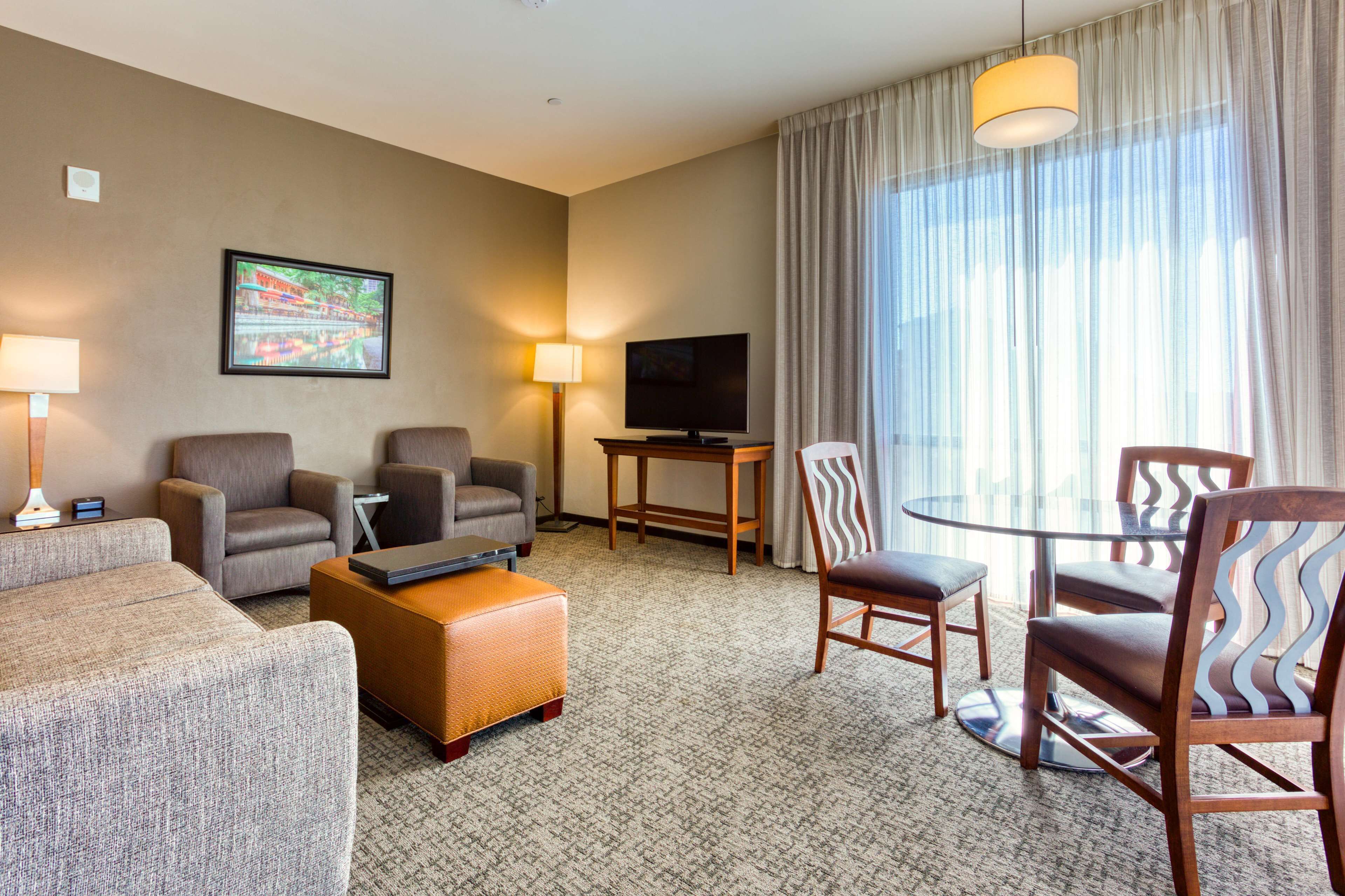 A hotel room with a sofa, two armchairs, a small table, a TV, and a dining area with chairs.