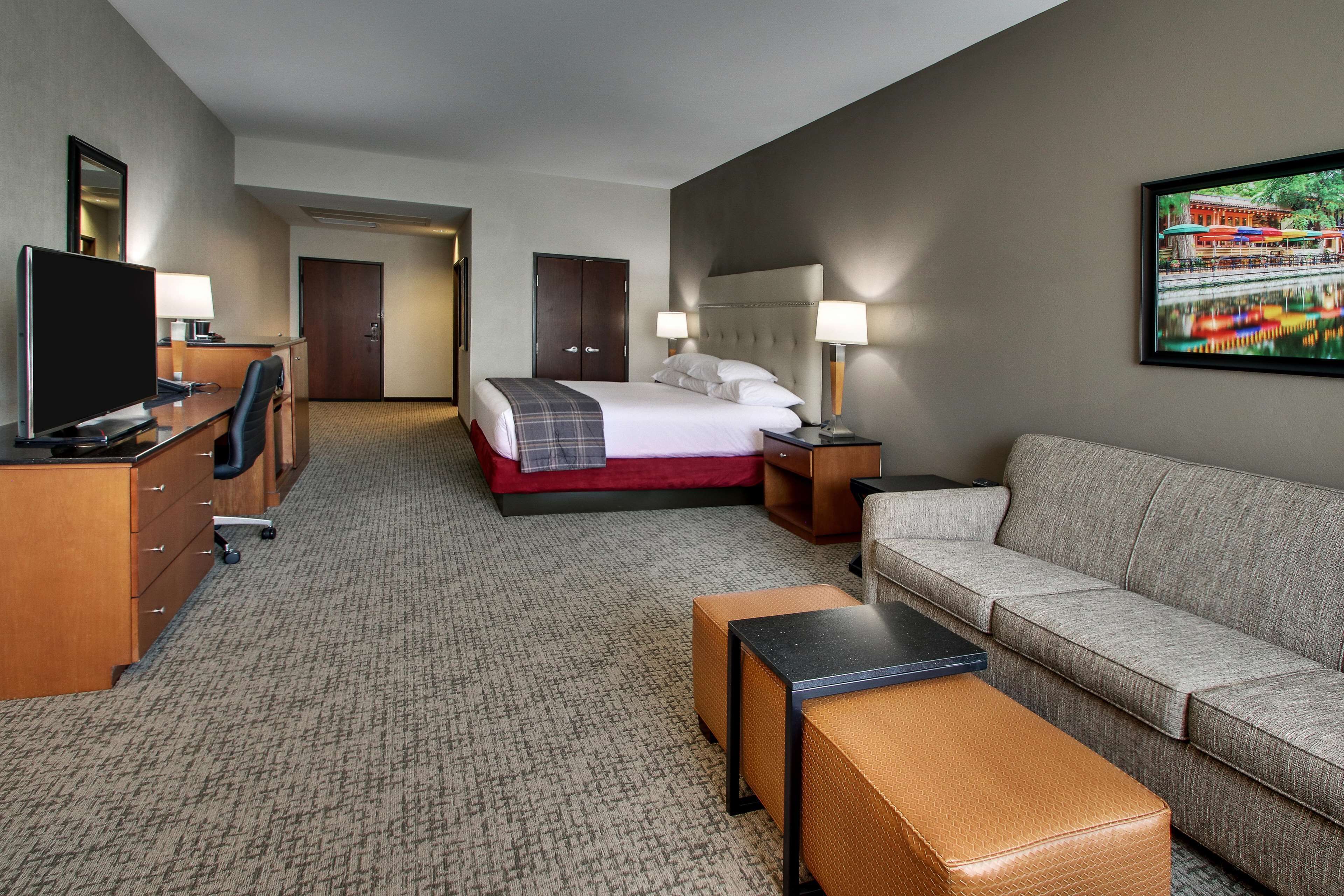 Ein Hotelzimmer mit Bett, Sofa, Schreibtisch mit Computer, Fernseher und einem Gemälde an der Wand.