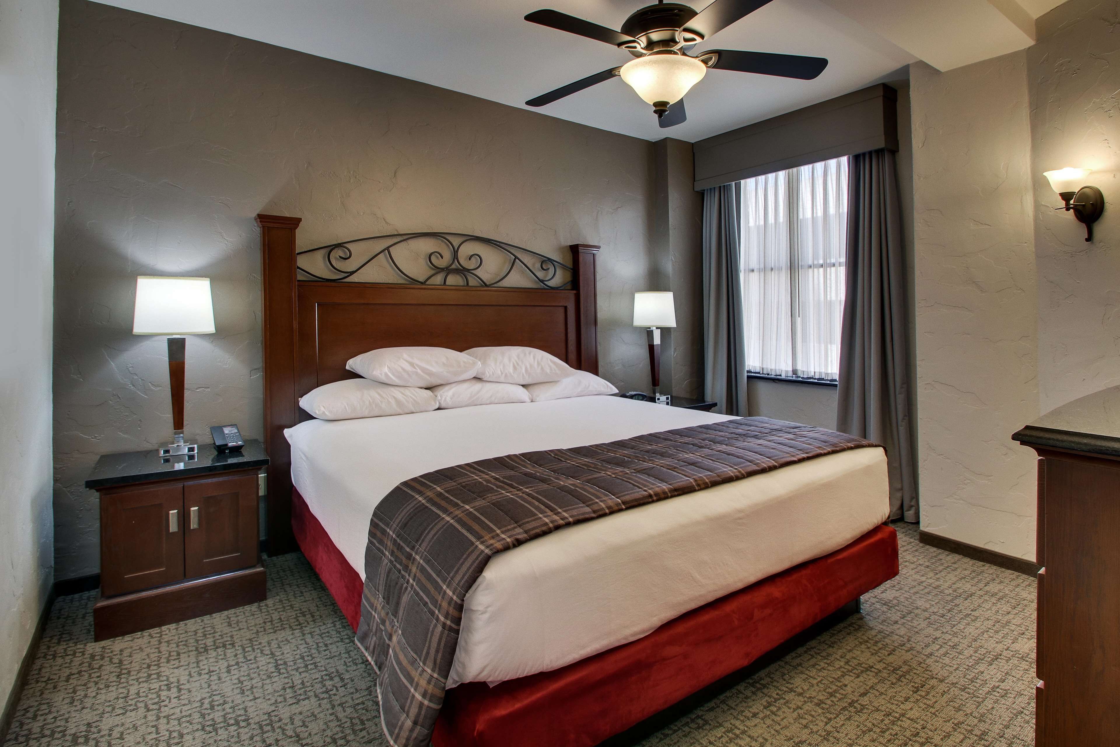 A hotel room with a large bed, two bedside lamps, a nightstand, a ceiling fan, and a window with curtains.