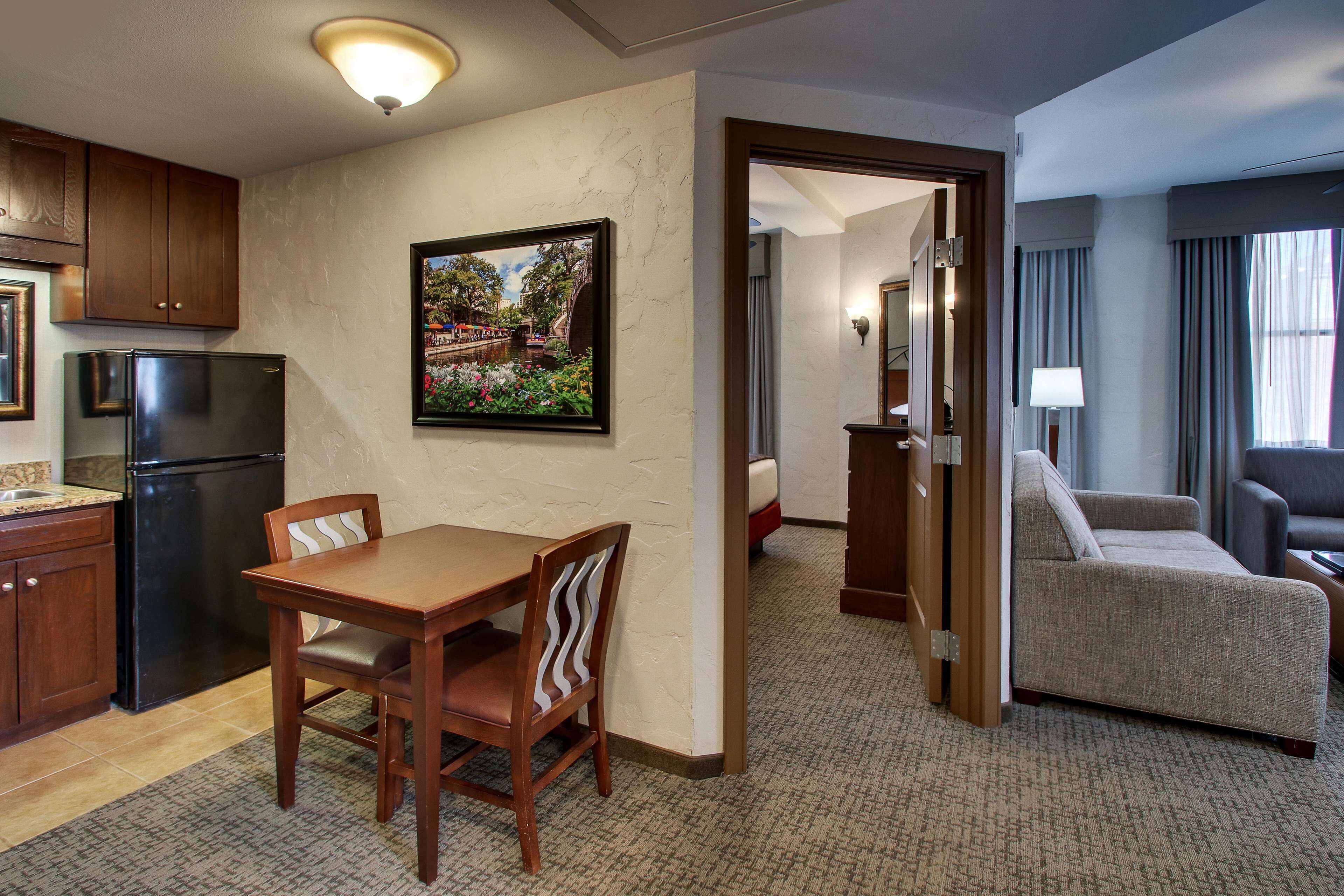 A hotel room with a dining area, a kitchenette, a living area with a sofa, and a door leading to another room.