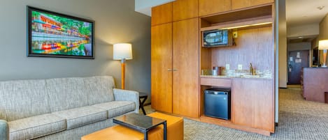 Una habitación de hotel con sofá, una mesita, un mueble con televisor y una zona de kitchenette.
