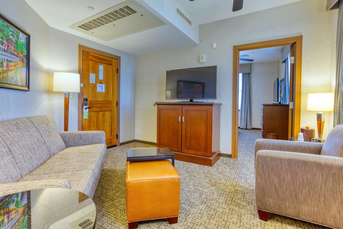 Une chambre d’hôtel équipée d’un canapé, d’un pouf, d’une télévision, d’une armoire en bois et d’un tableau accroché au mur.