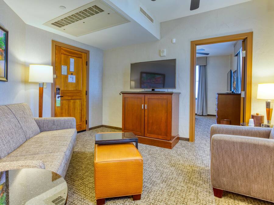 Ein Hotelzimmer mit Sofa, Hocker, Fernseher, Holzschrank und einem Gemälde an der Wand.