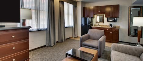 A hotel room with a TV, a sofa, a chair, a coffee table, a ceiling fan, a kitchen area with cabinets and a refrigerator, and a large window with curtains.
