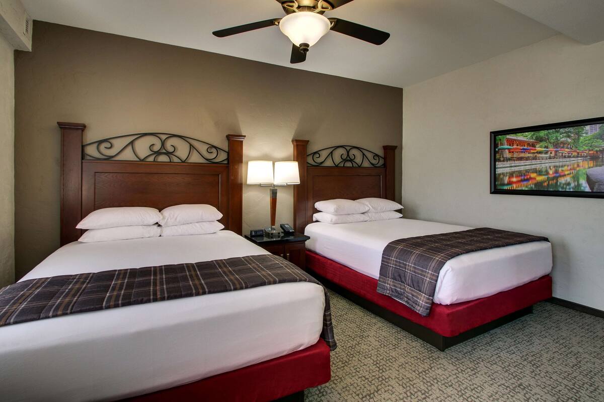 A hotel room with two beds, a ceiling fan, a nightstand with a lamp, and a framed picture on the wall.