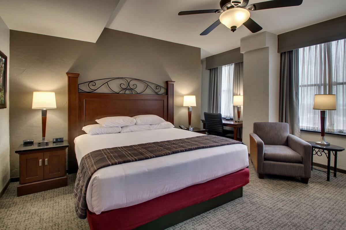 Une chambre d’hôtel avec un grand lit, deux tables de chevet, une chaise, un ventilateur de plafond et une fenêtre avec des rideaux.