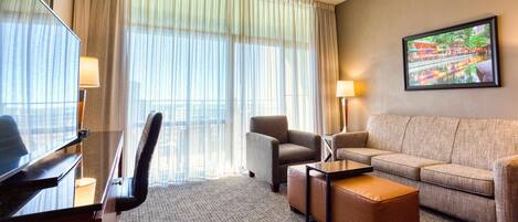 A hotel room with a large window, a sofa, a chair, a desk, and a television.