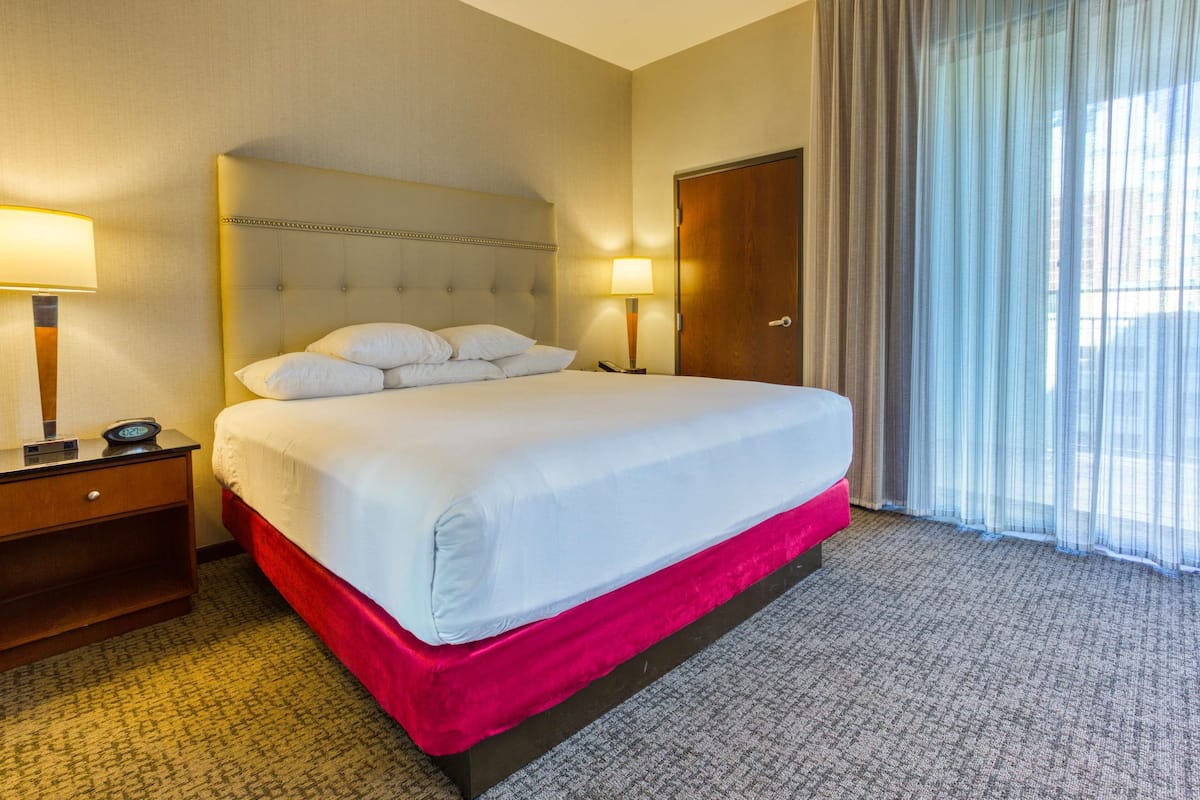 Une chambre d’hôtel avec un grand lit, une table de chevet avec une lampe, une porte en bois et une fenêtre avec des rideaux légers.