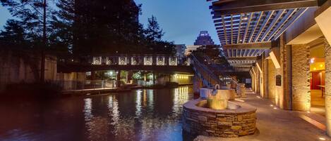 A riverside walkway with stone steps, a stone fountain, and a covered area with a metal roof.