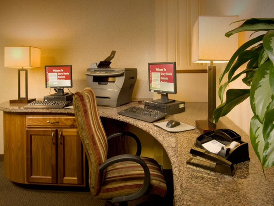 A hotel reception desk with two monitors displaying a welcome message, a desktop computer, a printer, a telephone, a chair, and a potted plant.