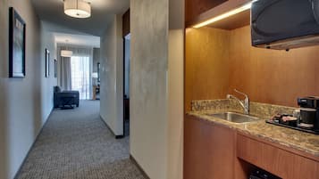 A hotel room with a kitchen area featuring a granite countertop, stainless steel sink, and a black dishwasher. A hallway with a carpeted floor and framed pictures on the wall is visible.