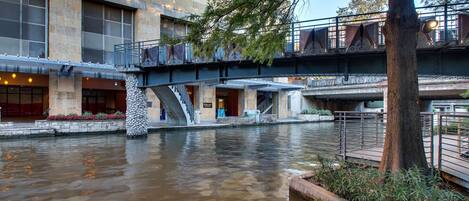 A riverside walkway with stone pillars, a bridge, and a building with large windows and a sign.