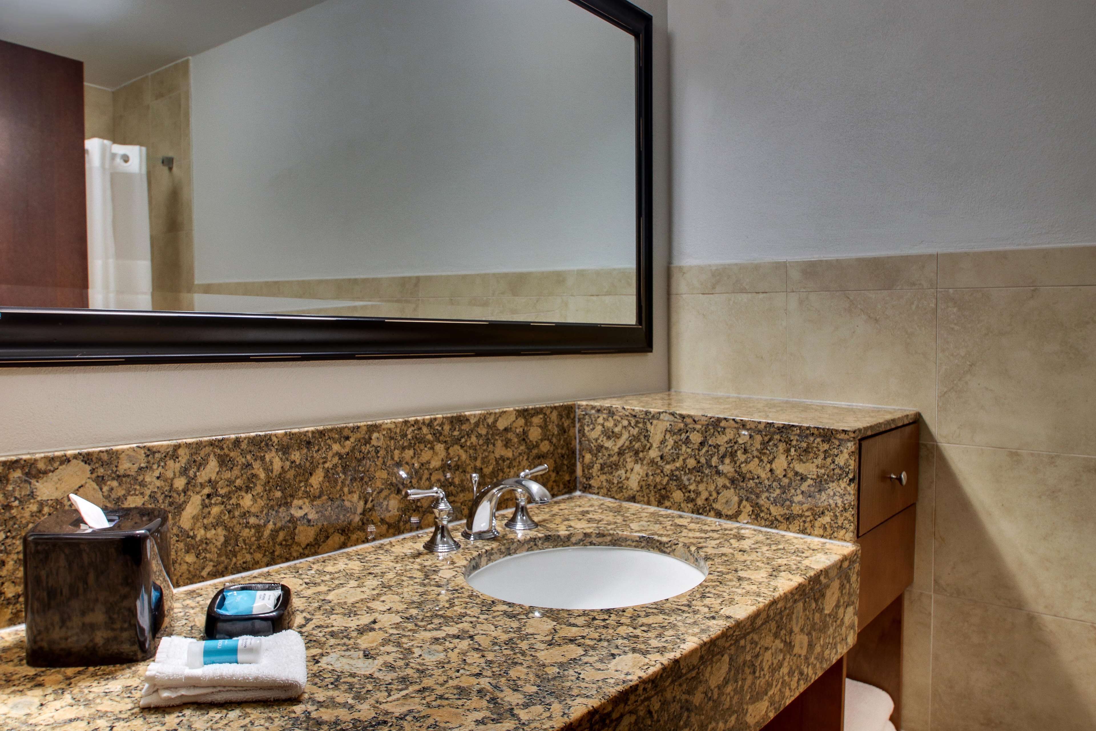 Un baño con encimera de granito, lavamanos, espejo y dispensador de toallas.