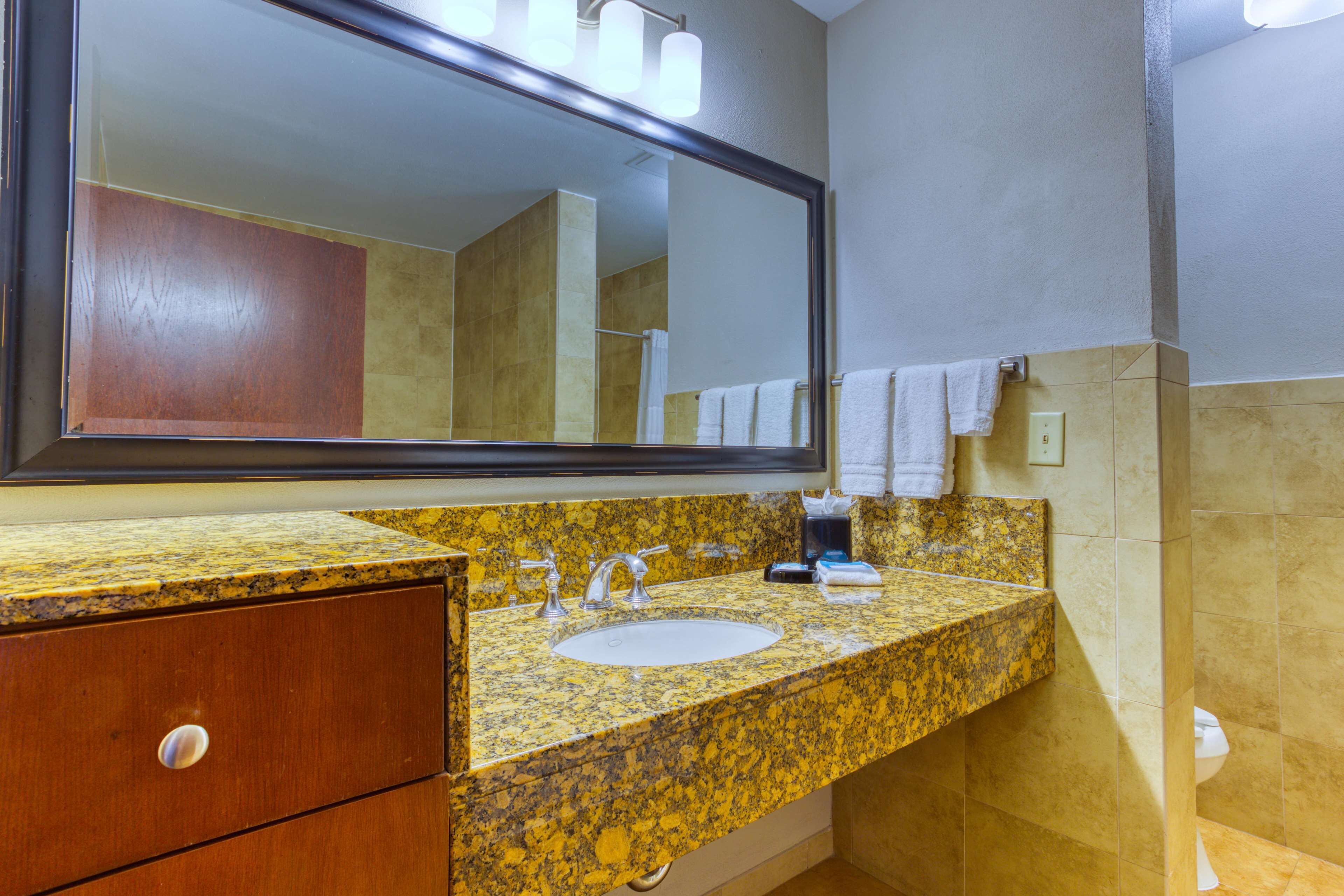 A bathroom with a granite countertop sink, a wooden cabinet, a large mirror, and beige tiled walls.