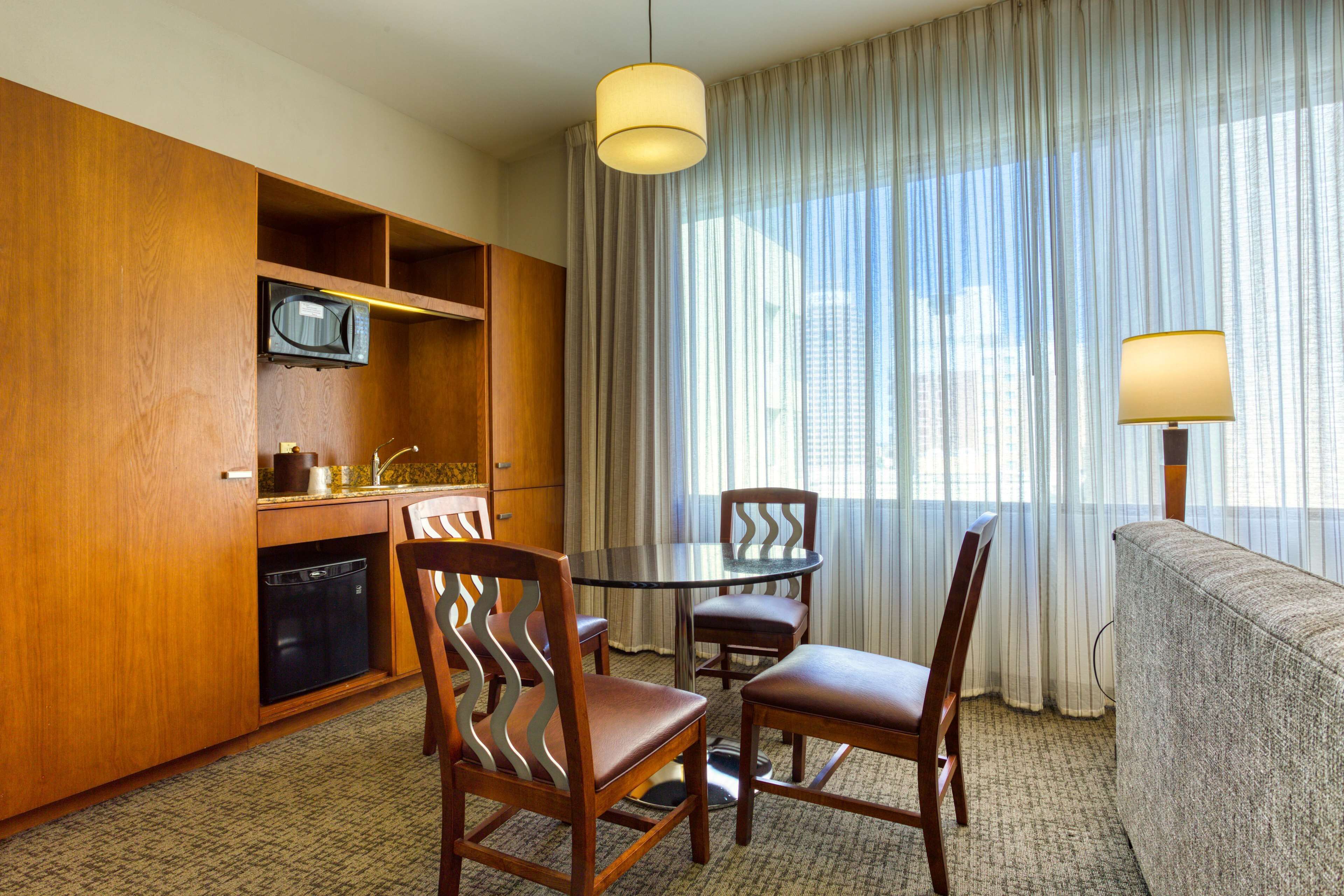 Une chambre d’hôtel compacte comprenant un coin cuisine, une table à manger avec quatre chaises, un canapé et une grande fenêtre avec des rideaux légers.