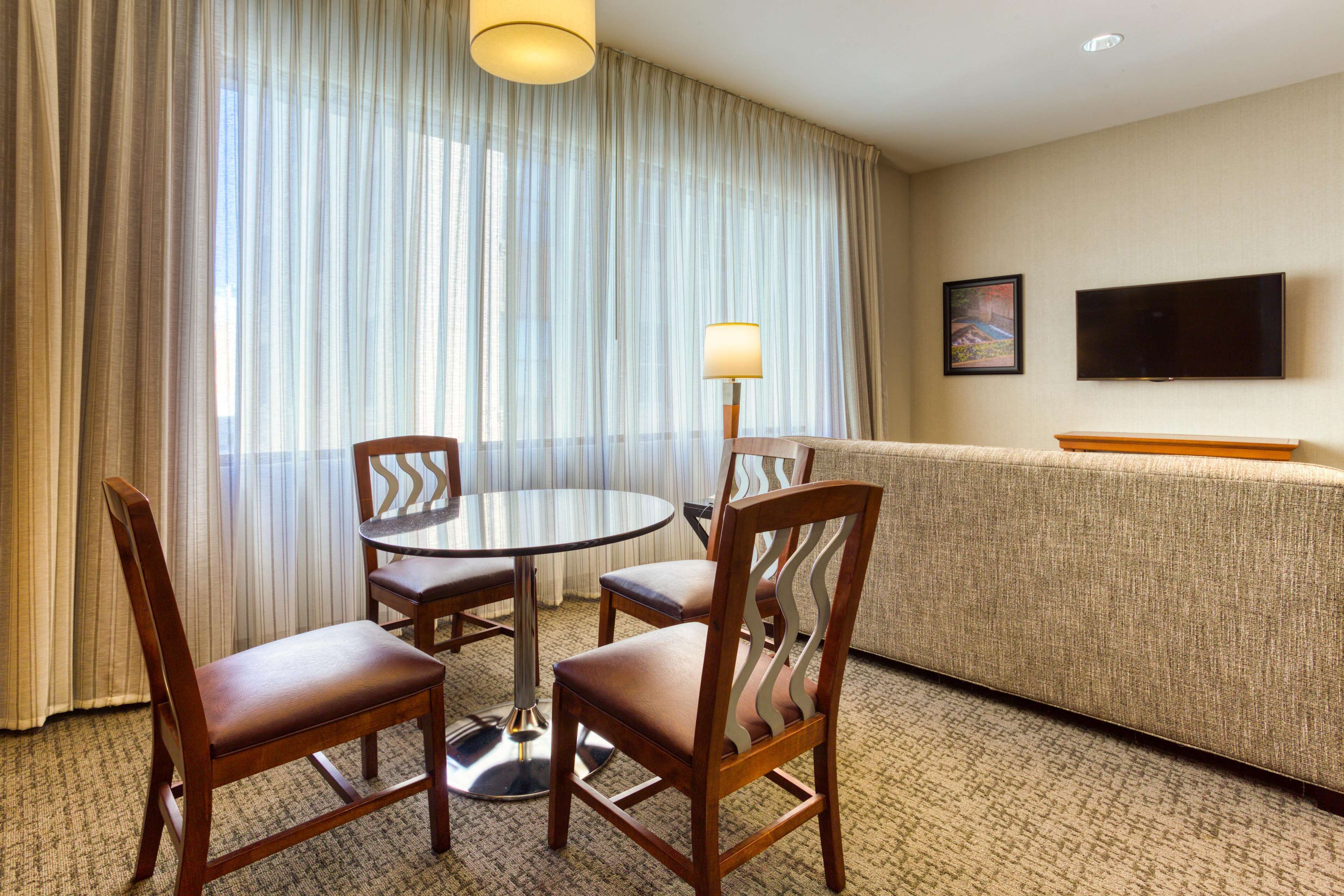 Habitación de hotel con una mesa redonda, cuatro sillas de madera, un sofá, un televisor montado en la pared y una ventana con cortinas traslúcidas.