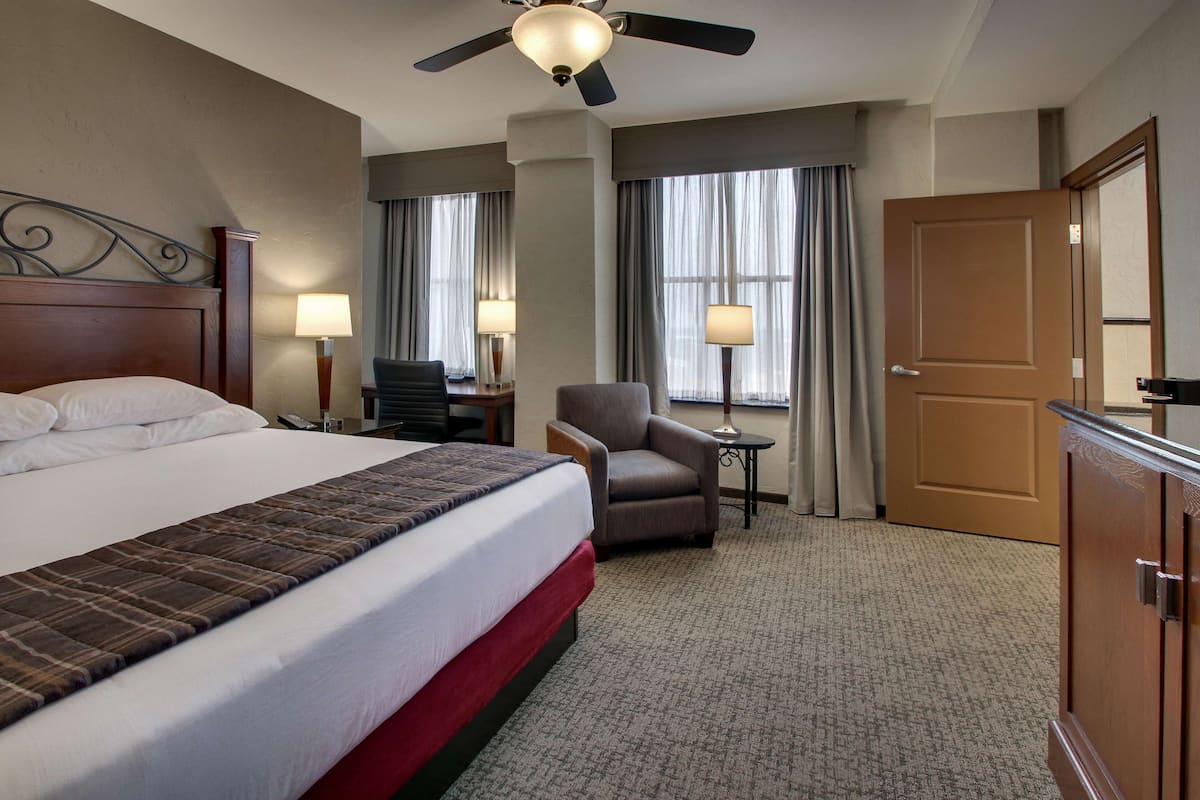 Une chambre d’hôtel avec un grand lit, un bureau avec une chaise, une petite table avec une lampe, un ventilateur de plafond et une porte.